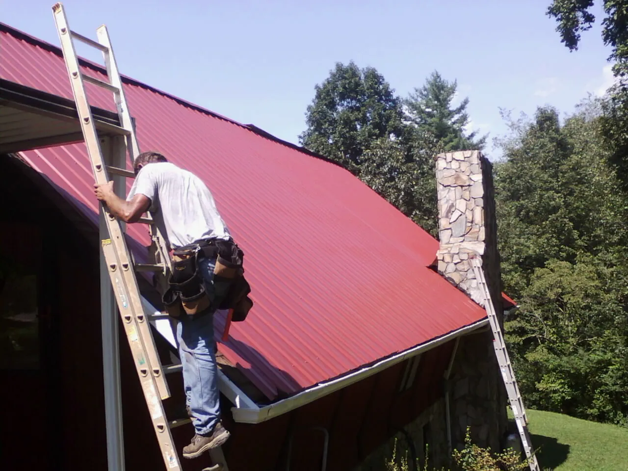 Roofer on a ladder beside a red metal roof and stone chimney — Soffit & Fascia Repair by Roofing Brothers in Purcell